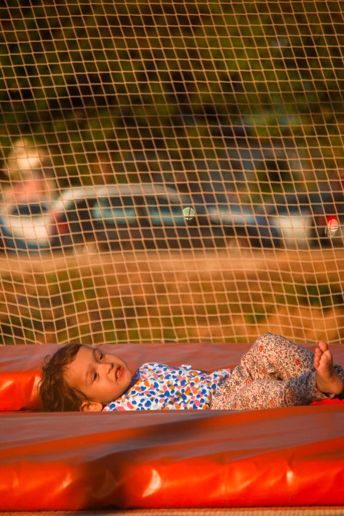 service-02 A young child in a colorful outfit enjoys lying on a trampoline outdoors, sunlit and carefree.